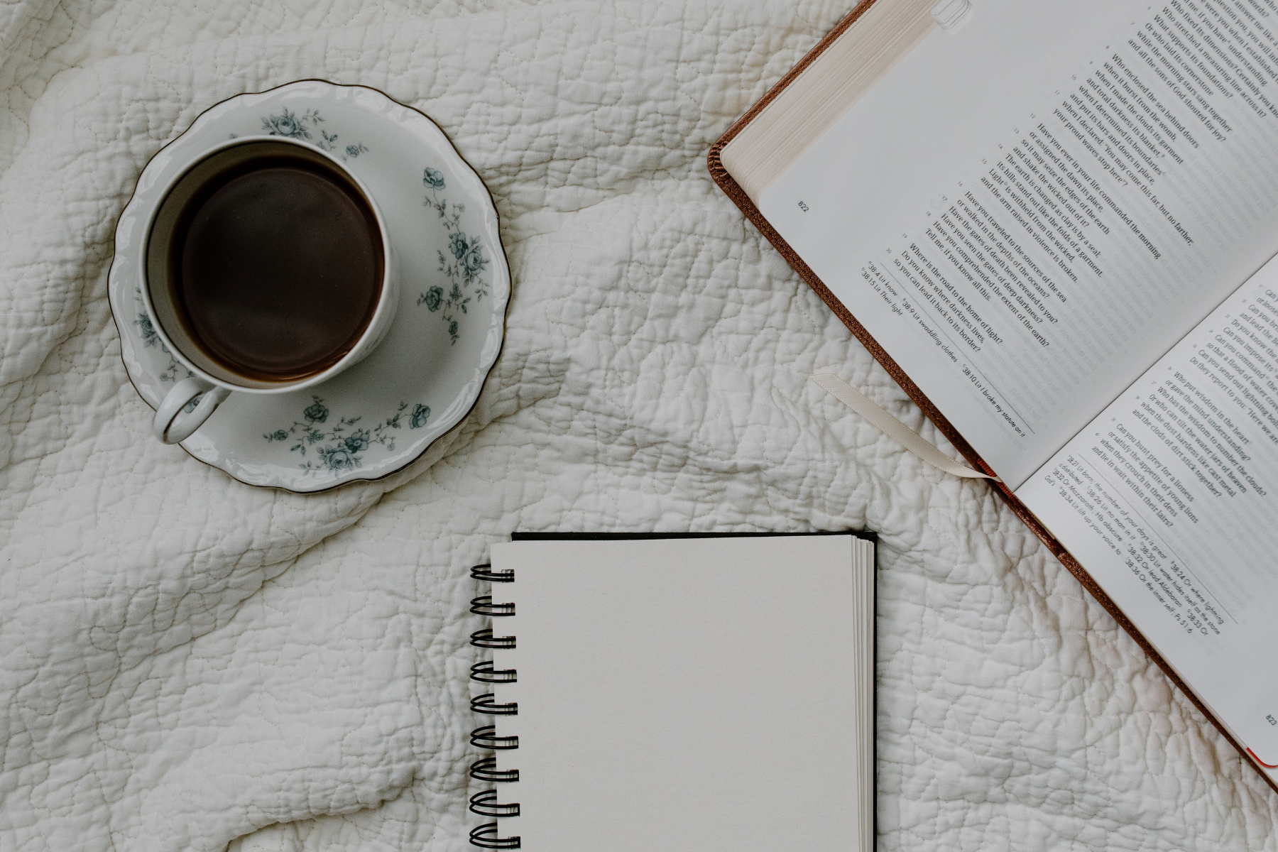 Cup of coffee on a plate next to an open bible and notebook on a white blanket